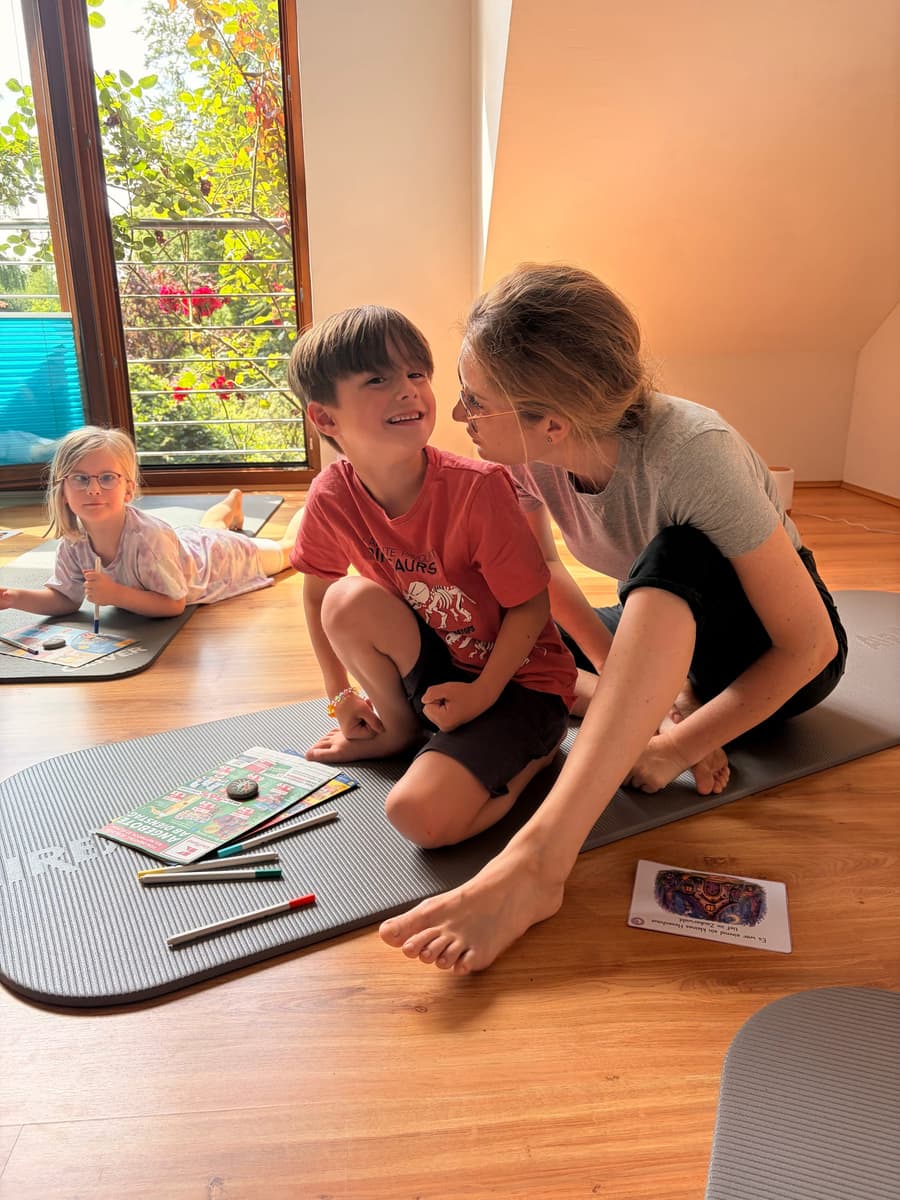 Ein Kind sitzt lachend auf einer Yogamatte und dreht sich zu einer erwachsenen Person um, die sich ihm zuwendet; daneben liegen Buntstifte, im Hintergrund ein sonniges Fenster.