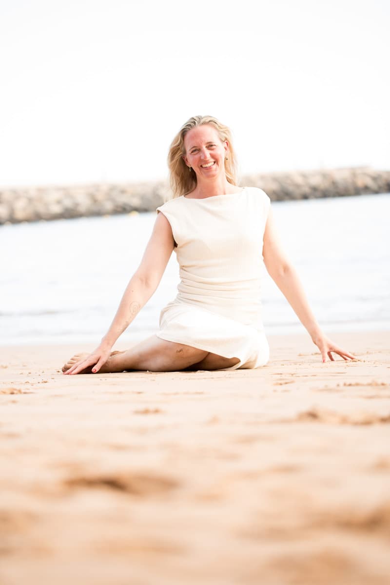 Janina sitzt im hellen Kleid im Sand, die Beine seitlich angewinkelt und die Hände abgestützt, und lacht offen in die Kamera; hinter ihr Wellen und eine Steinmole.