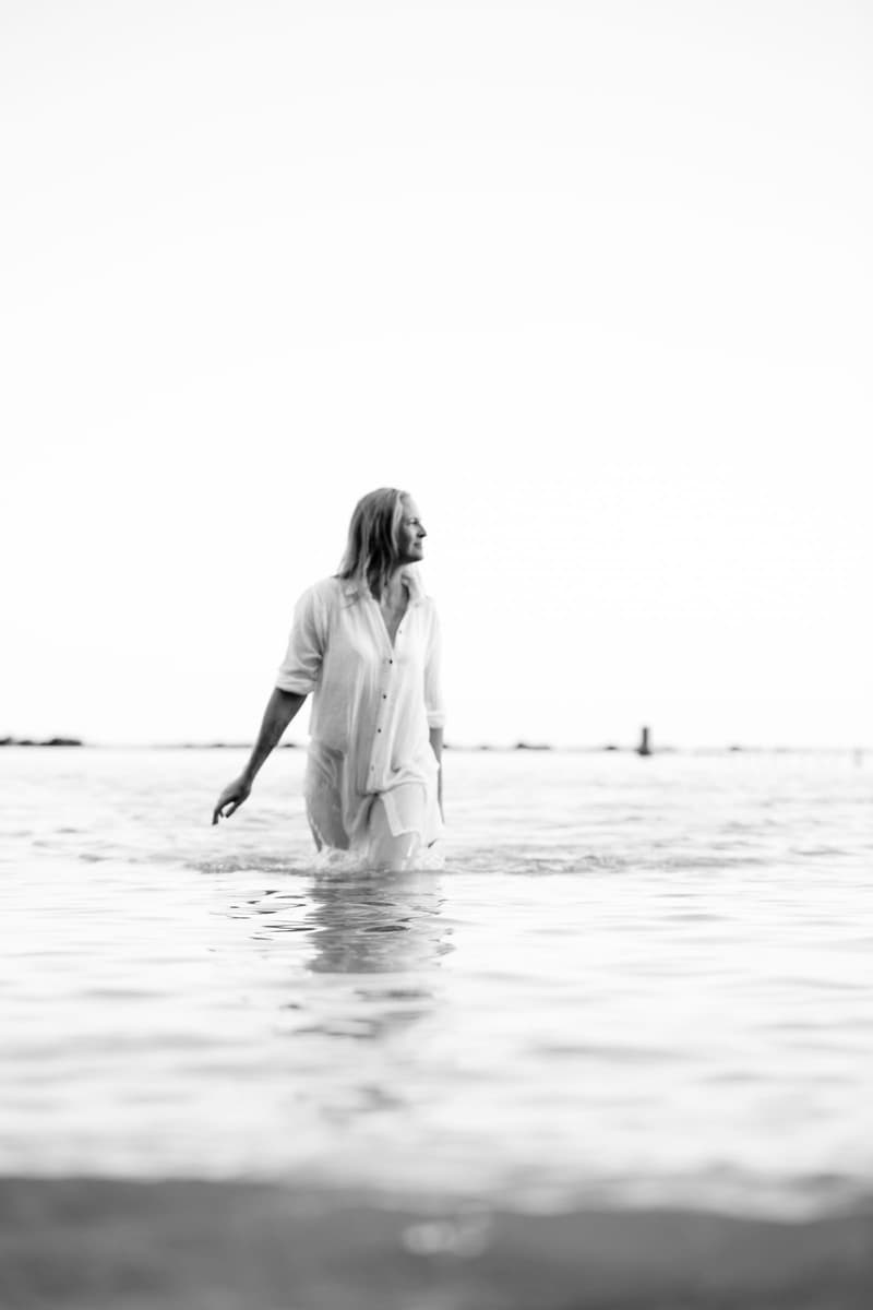 Schwarzweiß-Aufnahme: Janina steht in einem weißen Hemd im hüfthohen Wasser vor dem offenen Meer und blickt zur Seite.