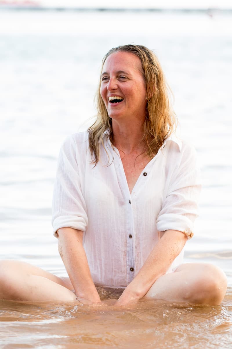 Janina sitzt im nassen weißen Hemd im flachen Wasser und lacht mit ganzem Gesicht zur Seite.