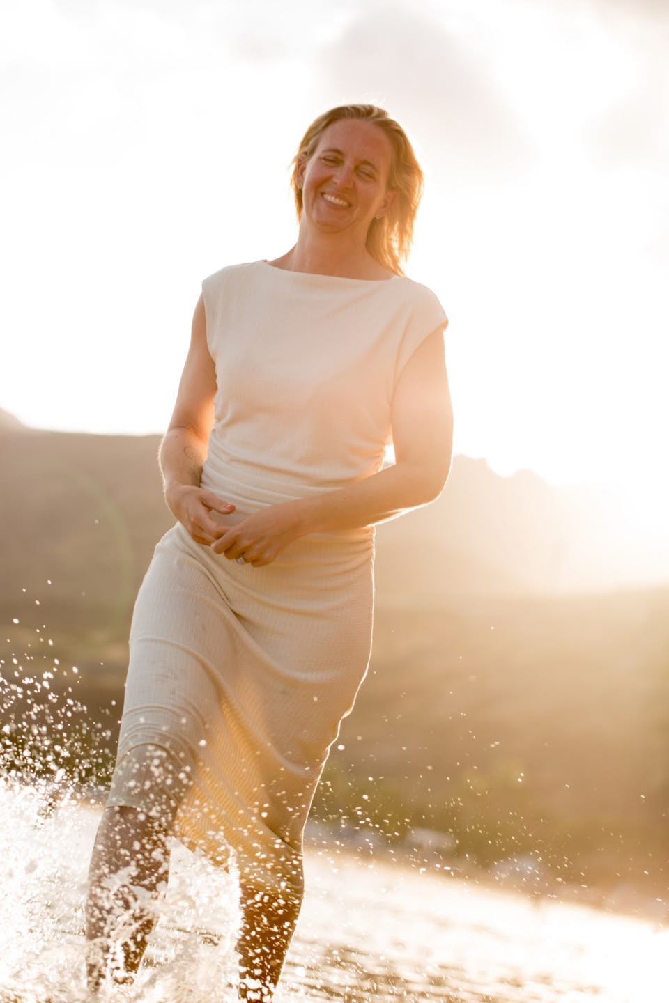 Janina läuft lachend durch knietiefes Wasser, Spritzer um die Beine; die tiefstehende Sonne steht als Gegenlicht hinter ihr.