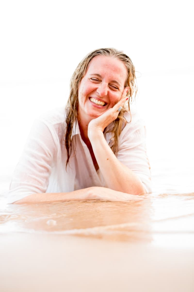 Janina liegt im weißen Hemd im flachen Wasser, den Kopf in die Hand gestützt, und lacht mit geschlossenen Augen.