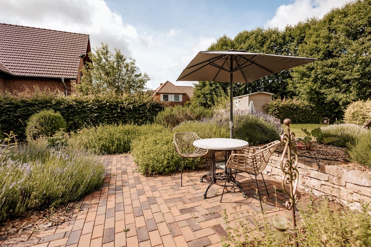 Der Garten des Okelmann’s: ein runder Tisch mit Korbsesseln unter einem Sonnenschirm, ringsum Lavendel, Buchsbaum und hohe Hecken vor dem Backsteinhaus.