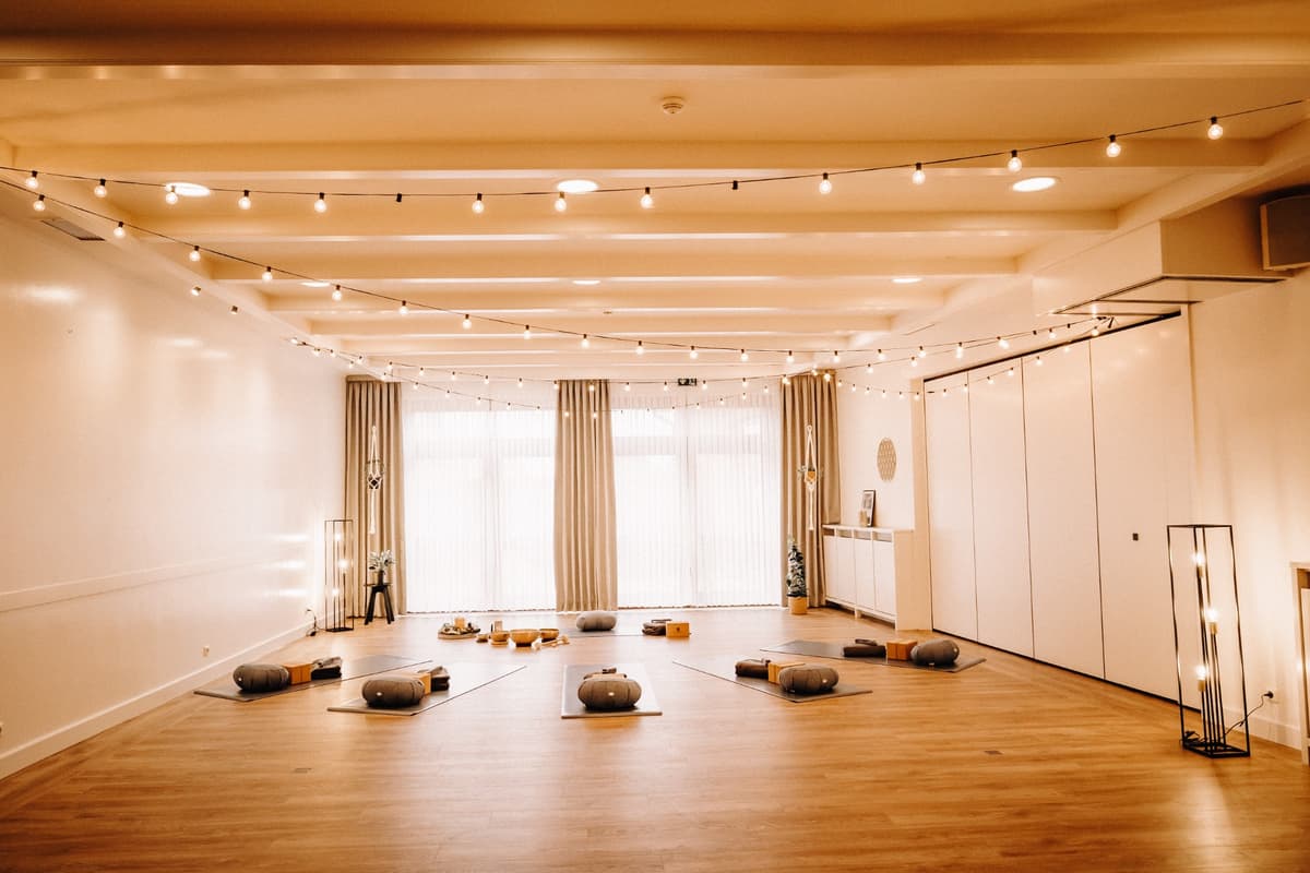 Der helle Yogaraum im Okelmann’s: Holzboden, ausgelegte Matten mit Kissen und Decken, Lichterketten unter der Decke und eine breite Fensterfront hinter Vorhängen.