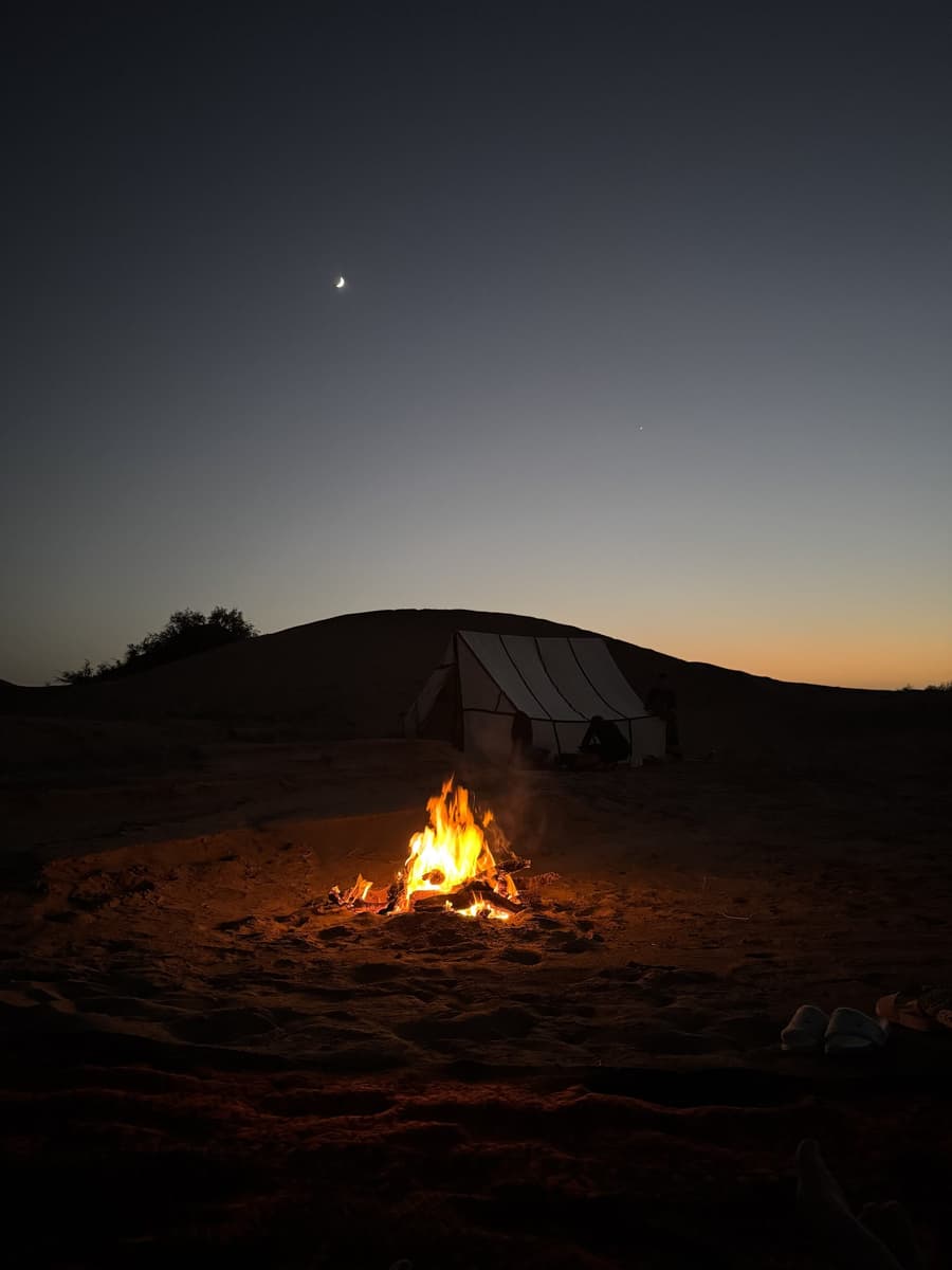 Abenddämmerung in der Sahara: über einer Düne stehen ein schmaler Mond und ein einzelner Stern, davor ein weißes Zelt und ein kleines Lagerfeuer im Sand.